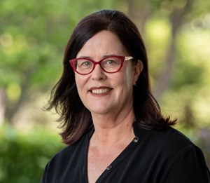 Headshot of Veronica Jeffrey - a women with red glasses, dark brown hair who is smiling against a leafy green background